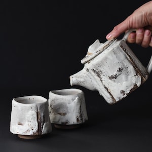 Può includere: Una teiera in ceramica bianca con una finitura rustica viene versata in due tazze in ceramica bianca abbinate. La teiera e le tazze hanno una superficie testurizzata e irregolare.