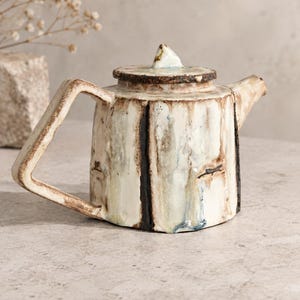 Op de afbeelding: Een keramische theepot met een getextureerd, gebroken wit glazuur met verticale bruine en zwarte strepen. De theepot heeft een geometrisch handvat, een korte tuit en een bijpassend deksel met een puntige knop, met een rustieke uitstraling.