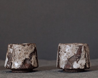May include: Two small, brown ceramic cups with white glaze. The cups have a rough, textured surface and are slightly asymmetrical.