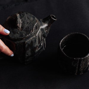 Op de afbeelding: Een zwarte theepot van keramiek met een getextureerd oppervlak en een bijpassende kop gevuld met donkere vloeistof. De theepot heeft een unieke, hoekige vorm.