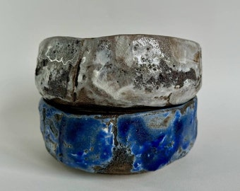 May include: Two ceramic bowls, one with a white and gray glaze and the other with a blue glaze. The bowls are stacked on top of each other.