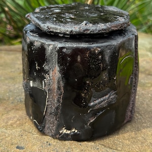 May include: A handmade, black ceramic jar with a lid. The jar has a cylindrical shape with a textured, rough surface. The lid is flat and slightly larger than the jar's opening. The glaze is glossy and reflects light, creating highlights and shadows.