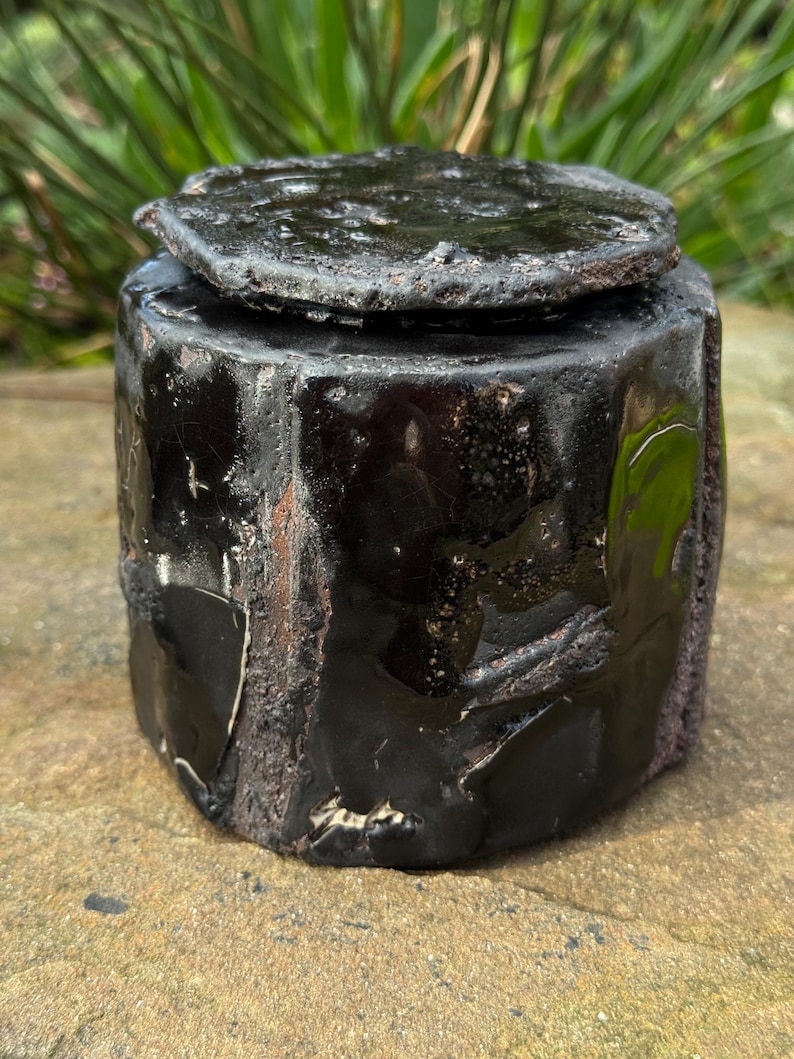 May include: A handmade, black ceramic jar with a lid. The jar has a cylindrical shape with a textured, rough surface. The lid is flat and slightly larger than the jar's opening. The glaze is glossy and reflects light, creating highlights and shadows.