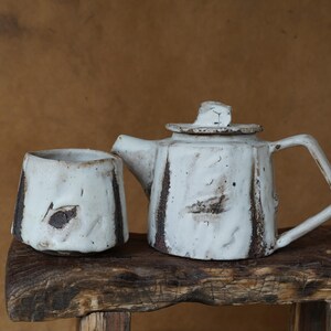 May include: A handmade ceramic teapot and cup set. The teapot has a lid and handle, and the cup is cylindrical. Both pieces are white with brown accents and sit on a rustic wooden surface.