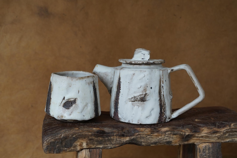May include: A handmade ceramic teapot and cup set. The teapot has a lid and handle, and the cup is cylindrical. Both pieces are white with brown accents and sit on a rustic wooden surface.