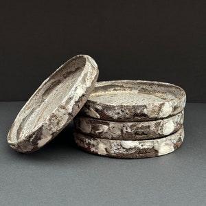 Peut inclure: Un ensemble de quatre assiettes en céramique avec un glaçage blanc et brun moucheté. Les assiettes sont empilées les unes sur les autres.