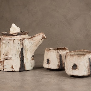 Op de afbeelding: Een keramische theepot met deksel en twee bijpassende kopjes, allemaal voorzien van een getextureerd, gebroken wit glazuur met donkerbruine, verticale penseelstreekaccenten en een ruwe, handgevormde uitstraling. De theepot heeft een brede greep.