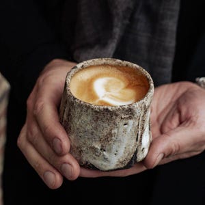 Op de afbeelding: Een close-up van een rustieke, bruin-witte keramische mok gevuld met een latte. De latte heeft een hartvormig ontwerp in het schuim.