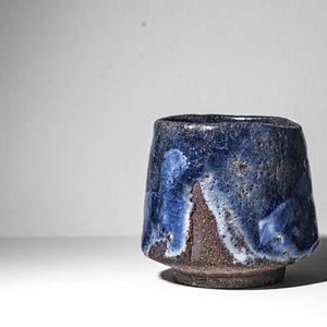 May include: A small, dark brown ceramic cup with a blue and white glaze. The glaze has a mottled, uneven appearance, creating a unique and artistic look.