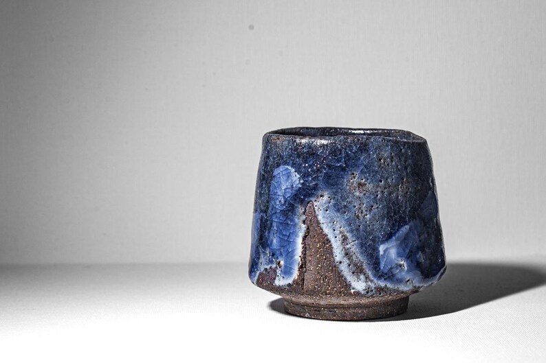 May include: A small, dark brown ceramic cup with a blue and white glaze. The glaze has a mottled, uneven appearance, creating a unique and artistic look.