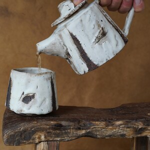 May include: A white ceramic teapot with a matching cup. The teapot is pouring liquid into the cup. Both pieces have dark brown accents and a textured, rustic appearance. The set is placed on a wooden surface.