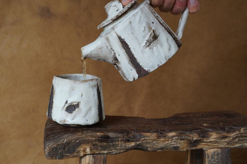May include: A white ceramic teapot with a matching cup. The teapot is pouring liquid into the cup. Both pieces have dark brown accents and a textured, rustic appearance. The set is placed on a wooden surface.