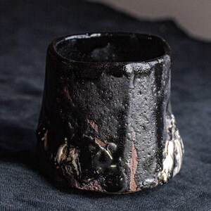 May include: A black ceramic cup with a textured, uneven surface. The cup has a wide, rounded base and a slightly flared rim. The surface of the cup is covered in small, dark brown spots and streaks.