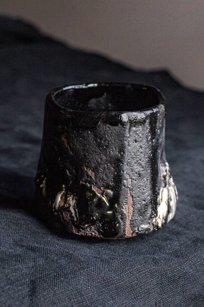 May include: A black ceramic cup with a textured, uneven surface. The cup has a wide, rounded base and a slightly flared rim. The surface of the cup is covered in small, dark brown spots and streaks.