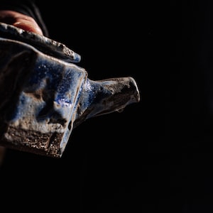 Könnte beinhalten: Eine handgefertigte, blaue Keramik-Teekanne mit strukturierter, rustikaler Optik. Der Ausguss ist sichtbar, und die Kanne wird vor einem dunklen Hintergrund gehalten. Die Teekanne hat eine einzigartige, handwerkliche Qualität.