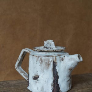 May include: A handmade ceramic teapot with a white glaze and dark brown accents. The teapot has a square body, a curved handle, and a lid with a small knob. The teapot is sitting on a wooden surface.