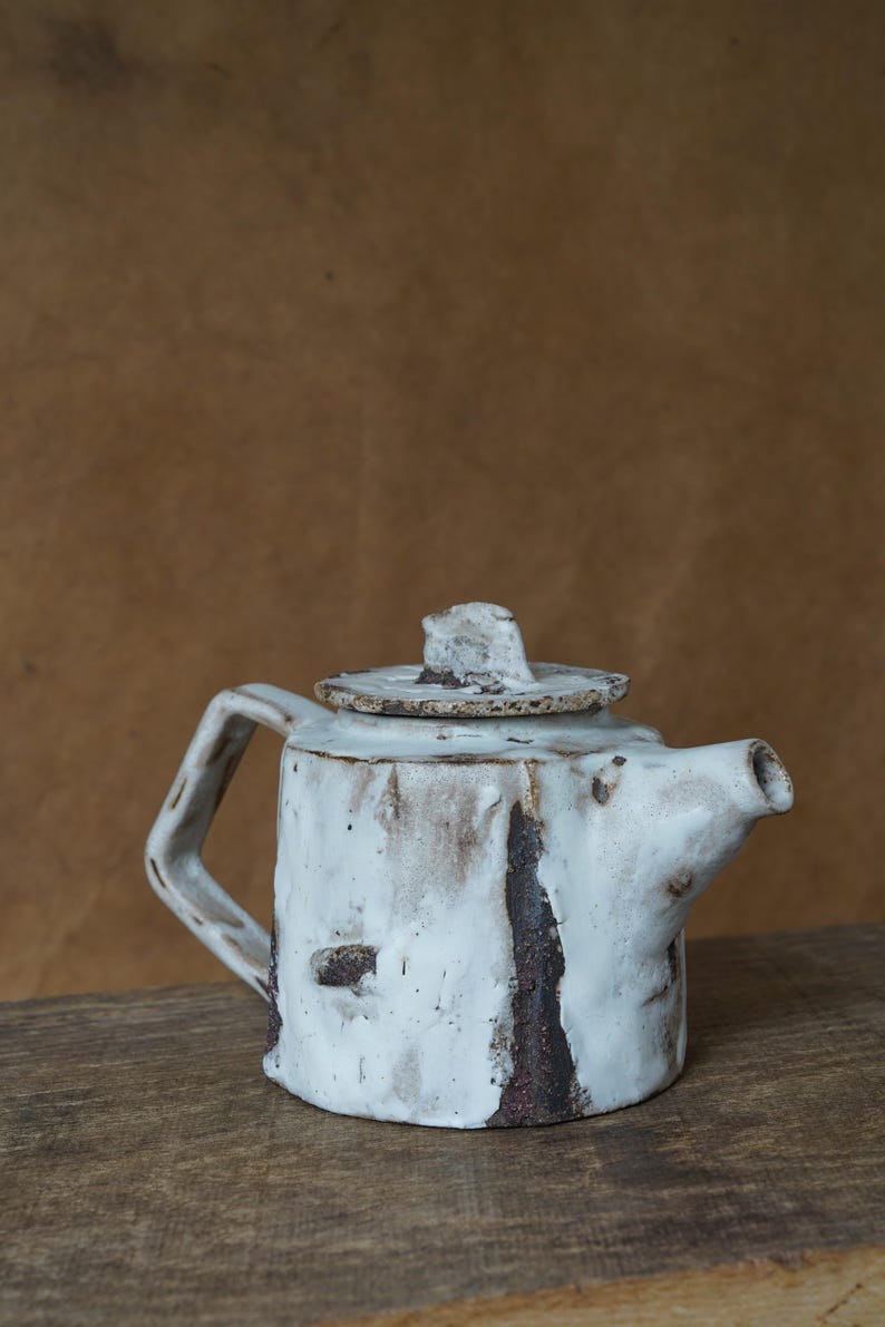 May include: A handmade ceramic teapot with a white glaze and dark brown accents. The teapot has a square body, a curved handle, and a lid with a small knob. The teapot is sitting on a wooden surface.