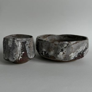 Op de afbeelding: Een set van twee keramische kommen, een klein en een groot, met een rustieke, textuurrijke afwerking. De kommen zijn grijs en wit gevlekt met hints van bruin en rood.