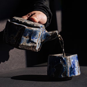 Puede incluir: Un juego de tetera y taza de cerámica azul con una apariencia texturizada y rústica. La tetera está vertiendo líquido en la taza. El juego tiene forma cuadrada y un esmalte azul oscuro con acentos marrones. La imagen está sobre un fondo oscuro.