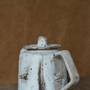 May include: A handmade white ceramic teapot with a lid and handle. The teapot has a rustic, textured appearance with brown and black accents. The design is simple and functional, suitable for serving tea.