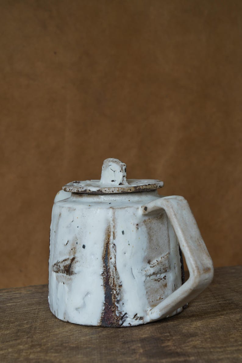 May include: A handmade white ceramic teapot with a lid and handle. The teapot has a rustic, textured appearance with brown and black accents. The design is simple and functional, suitable for serving tea.