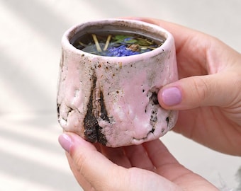 May include: A small, hand-formed ceramic cup with a textured, uneven surface glazed in pale pink and dark brown. The cup has a thick, irregular rim and contains a liquid with visible herbal tea leaves and a single blue flower petal floating inside.