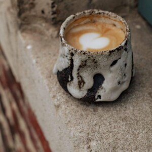 May include: A close-up of a cup of coffee with a heart-shaped design in the foam. The cup is made of a white ceramic with a brown glaze and has a rustic, handmade look.