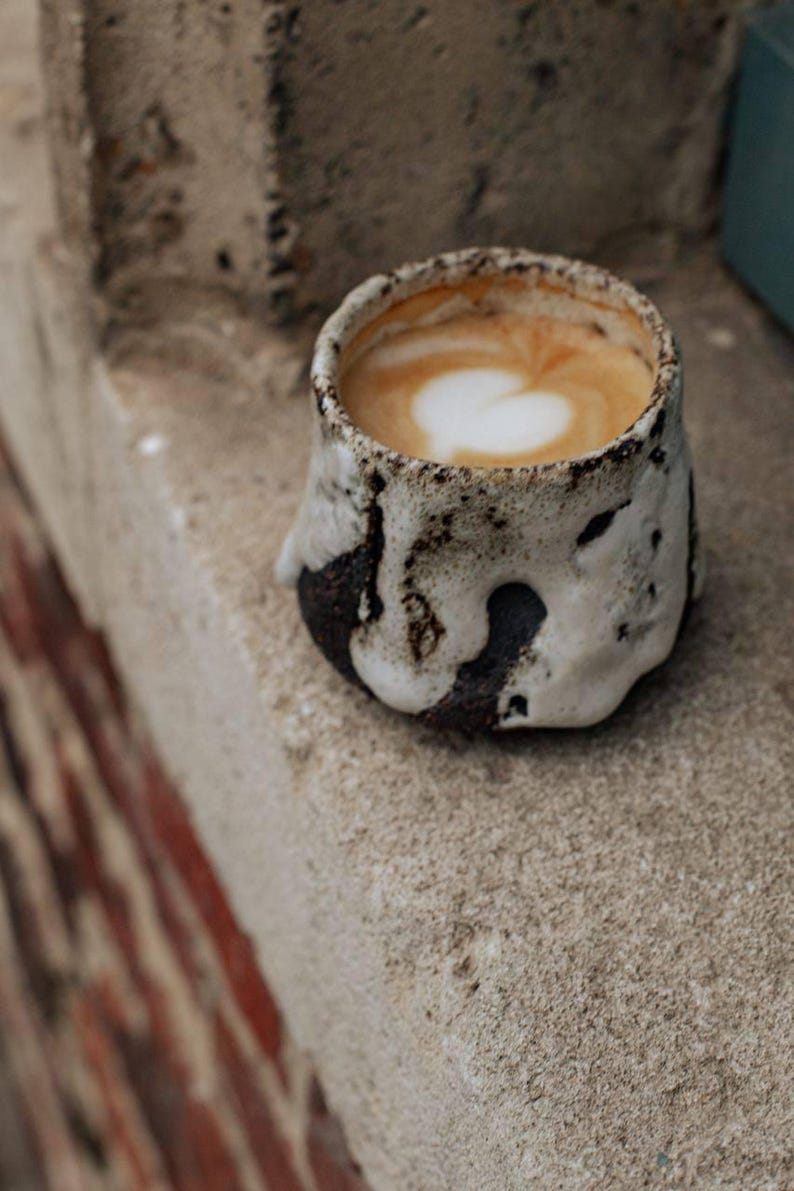 May include: A close-up of a cup of coffee with a heart-shaped design in the foam. The cup is made of a white ceramic with a brown glaze and has a rustic, handmade look.