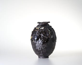 May include: A dark, textured, oval-shaped vase with a glossy black finish. The surface features irregular, raised bumps and small, light-colored, translucent flecks embedded within the dark glaze, leading to a narrow, circular opening at the top.