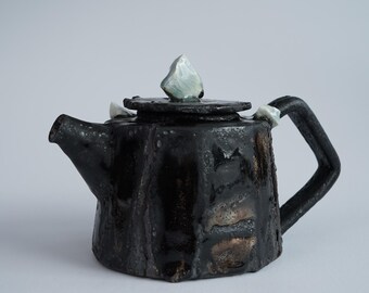 May include: A handcrafted black ceramic teapot with a unique, textured surface. The teapot features a geometric shape, a curved spout, and a handle. The lid is adorned with a light-colored, crystalline accent. A decorative piece for the kitchen.