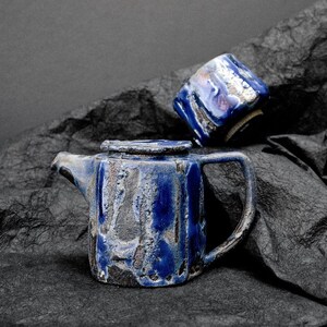Peut inclure: Une théière en céramique bleue et grise et une tasse assortie. La théière a une surface texturée et un bec verseur. La tasse est ronde et a une anse.