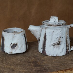 May include: A handmade ceramic tea set featuring a white teapot with a lid and a matching cup. The set has a rustic, textured appearance with brown accents. The teapot has a handle and spout, and the cup is cylindrical.