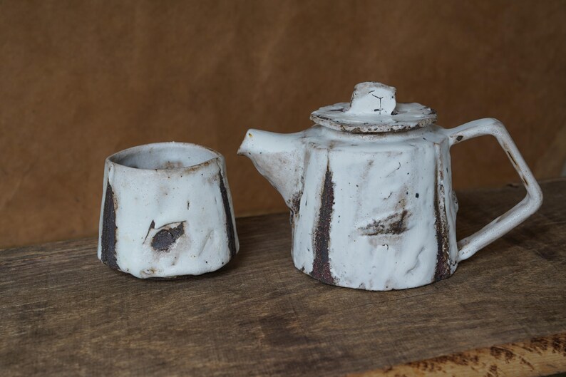 May include: A handmade ceramic tea set featuring a white teapot with a lid and a matching cup. The set has a rustic, textured appearance with brown accents. The teapot has a handle and spout, and the cup is cylindrical.