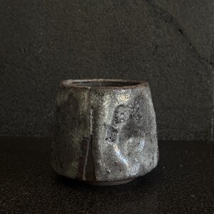 May include: A dark gray ceramic cup with a textured surface and a light gray glaze. The cup has a wide base and a slightly flared rim.