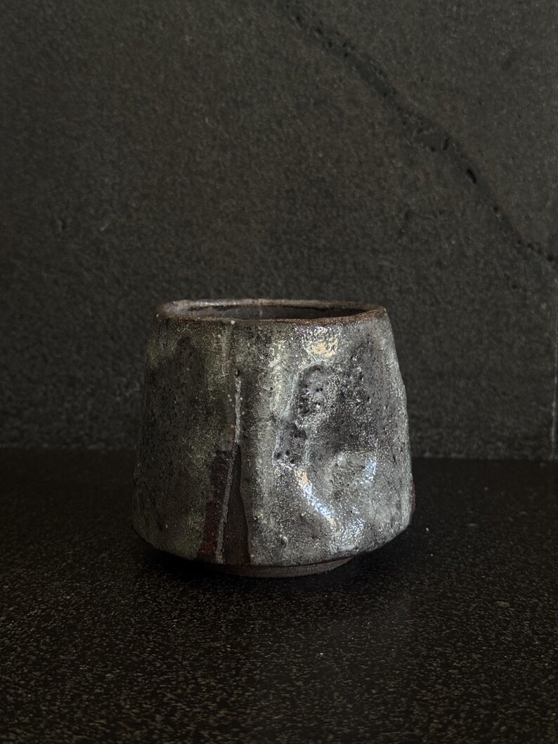 May include: A dark gray ceramic cup with a textured surface and a light gray glaze. The cup has a wide base and a slightly flared rim.