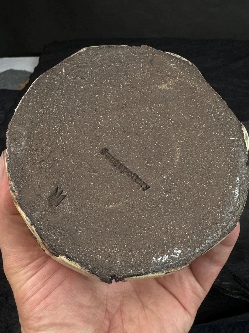 May include: A handmade ceramic pottery piece with a textured, dark brown base. The bottom of the pottery has a rough, unglazed surface with a stamped maker's mark. The pottery is held in a person's hand.