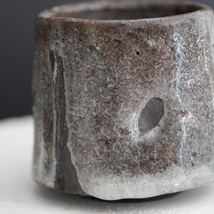 May include: A dark brown ceramic cup with a white glaze that has a textured, bubbly surface. The glaze has run down the sides of the cup, creating a unique and rustic look.