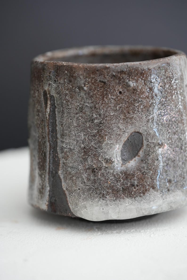 May include: A dark brown ceramic cup with a white glaze that has a textured, bubbly surface. The glaze has run down the sides of the cup, creating a unique and rustic look.