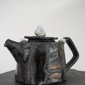 Peut inclure: Théière en céramique noire faite à la main avec une surface texturée. La théière a une poignée carrée et un couvercle orné d'une pierre bleu clair. Le bec verseur est incliné, et le design général est rustique et unique.