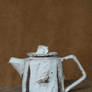 Puede incluir: Tetera de cerámica blanca hecha a mano con asa cuadrada y tapa. La tetera tiene una superficie texturizada con detalles marrones y un pico. La tetera está sobre una superficie de madera.