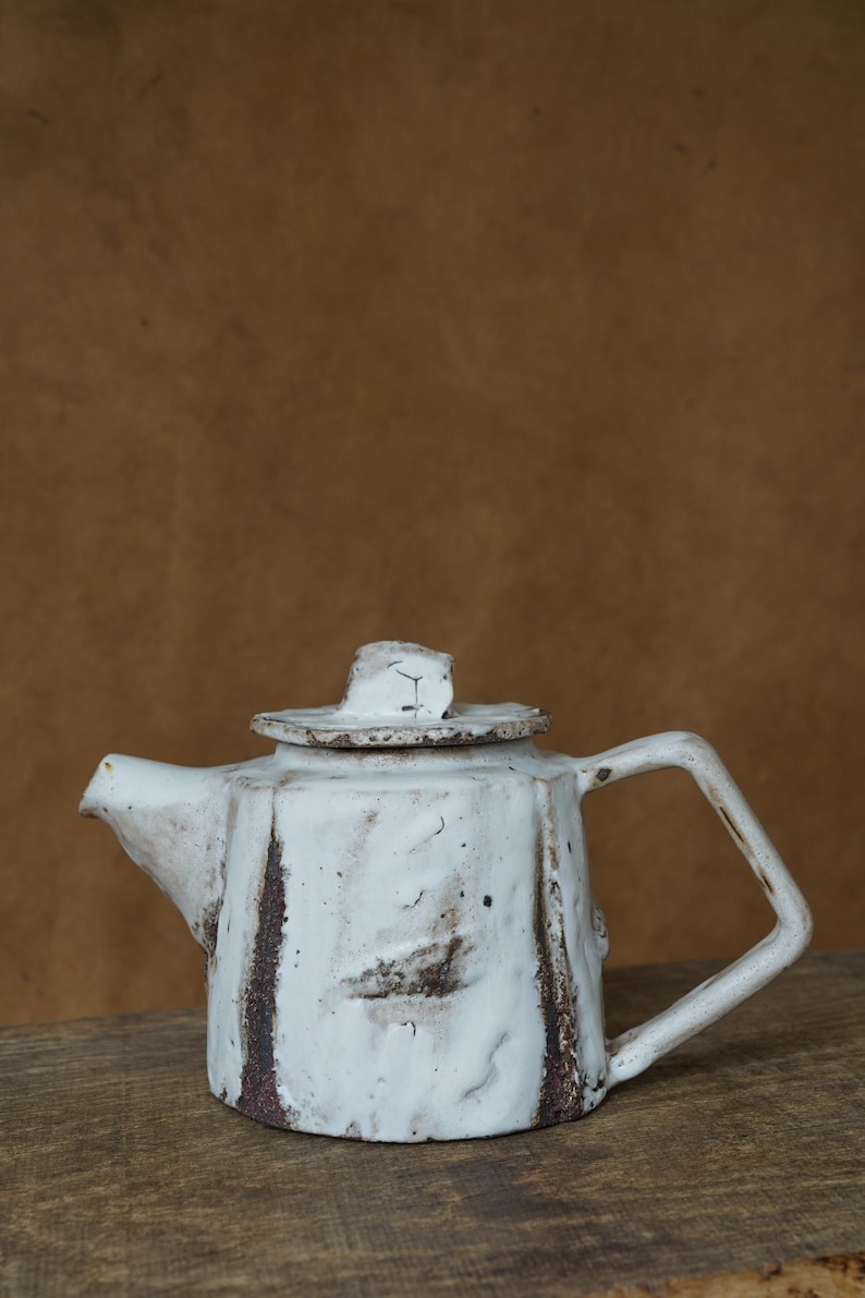 May include: A handmade white ceramic teapot with a square handle and a lid. The teapot has a textured surface with brown accents and a spout. The teapot is sitting on a wooden surface.