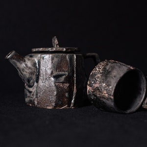 May include: A dark grey ceramic teapot and cup with a textured, rustic finish. The teapot has a lid and a spout, while the cup has a wide rim.