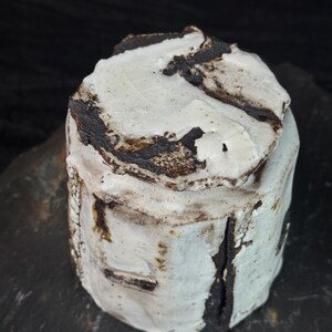 May include: A handmade ceramic jar with a lid. The jar is cylindrical with a rough, textured surface. It is primarily white with dark brown and black accents. The lid has a similar design. The jar is sitting on a dark surface.