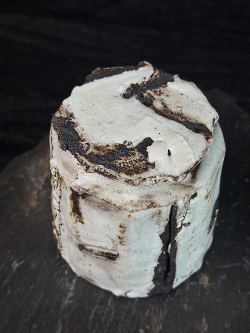 May include: A handmade ceramic jar with a lid. The jar is cylindrical with a rough, textured surface. It is primarily white with dark brown and black accents. The lid has a similar design. The jar is sitting on a dark surface.