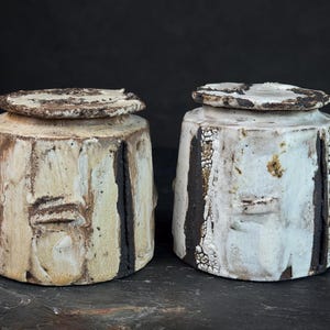 May include: Two handmade ceramic jars with lids. The jars are cylindrical with a rough, textured surface in off-white and cream tones. Dark brown vertical stripes and accents add contrast. The lids are circular and match the jars' rustic aesthetic.