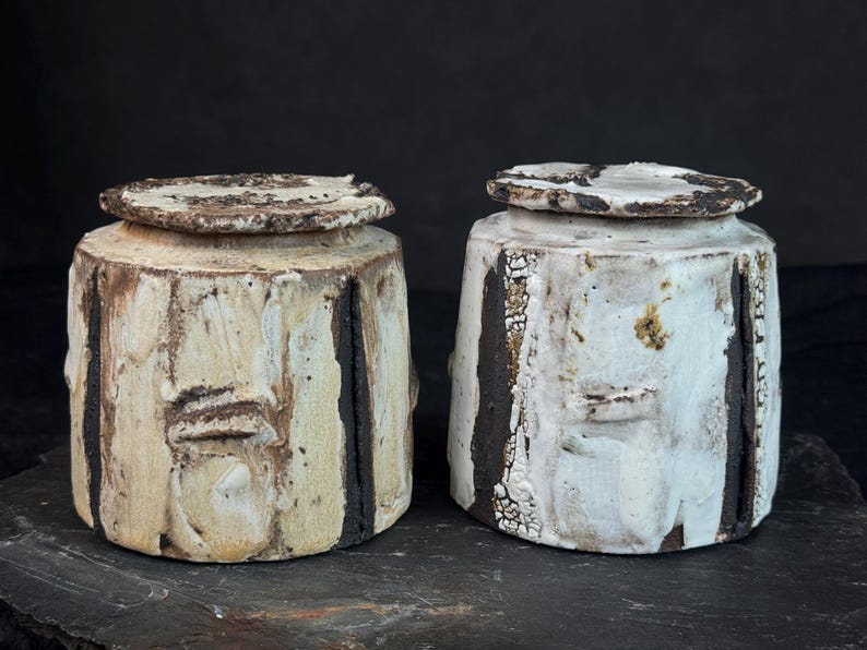 May include: Two handmade ceramic jars with lids. The jars are cylindrical with a rough, textured surface in off-white and cream tones. Dark brown vertical stripes and accents add contrast. The lids are circular and match the jars' rustic aesthetic.