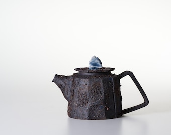 May include: A dark, textured, charcoal-colored teapot with a faceted, rugged exterior surface. The teapot features a geometric handle, a short spout, and a flat lid topped with a small, light blue, crystalline stone knob as a decorative handle.