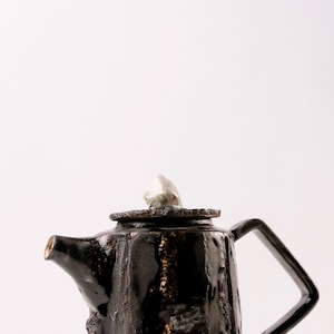 Può includere: Teiera in ceramica nera fatta a mano con una superficie strutturata e un design unico del manico. La teiera presenta un piccolo accento di colore chiaro sul coperchio. Il beccuccio è angolato e il design è moderno e artistico.