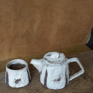 May include: A handmade white ceramic teapot and matching cup. The teapot has a square handle and a lid with a small knob. Both pieces have dark brown accents and sit on a wooden surface. The cup contains a dark liquid.