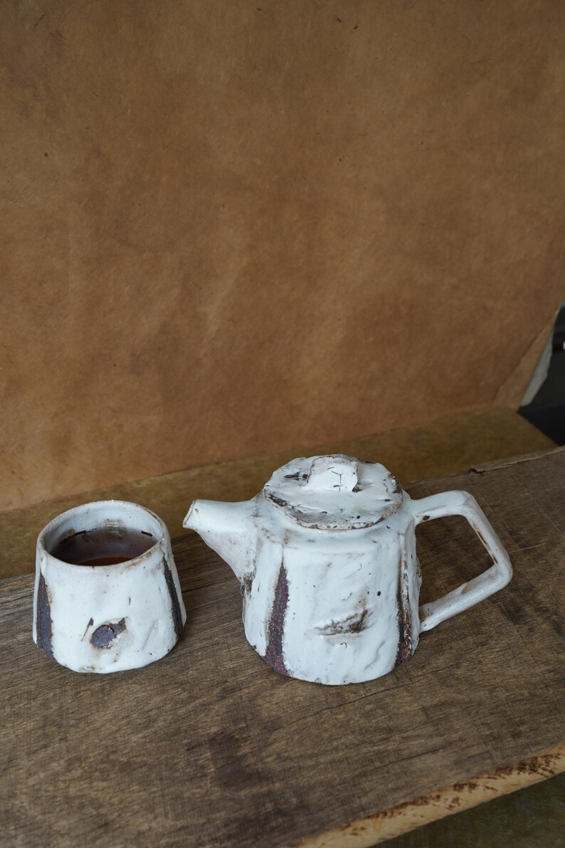 May include: A handmade white ceramic teapot and matching cup. The teapot has a square handle and a lid with a small knob. Both pieces have dark brown accents and sit on a wooden surface. The cup contains a dark liquid.